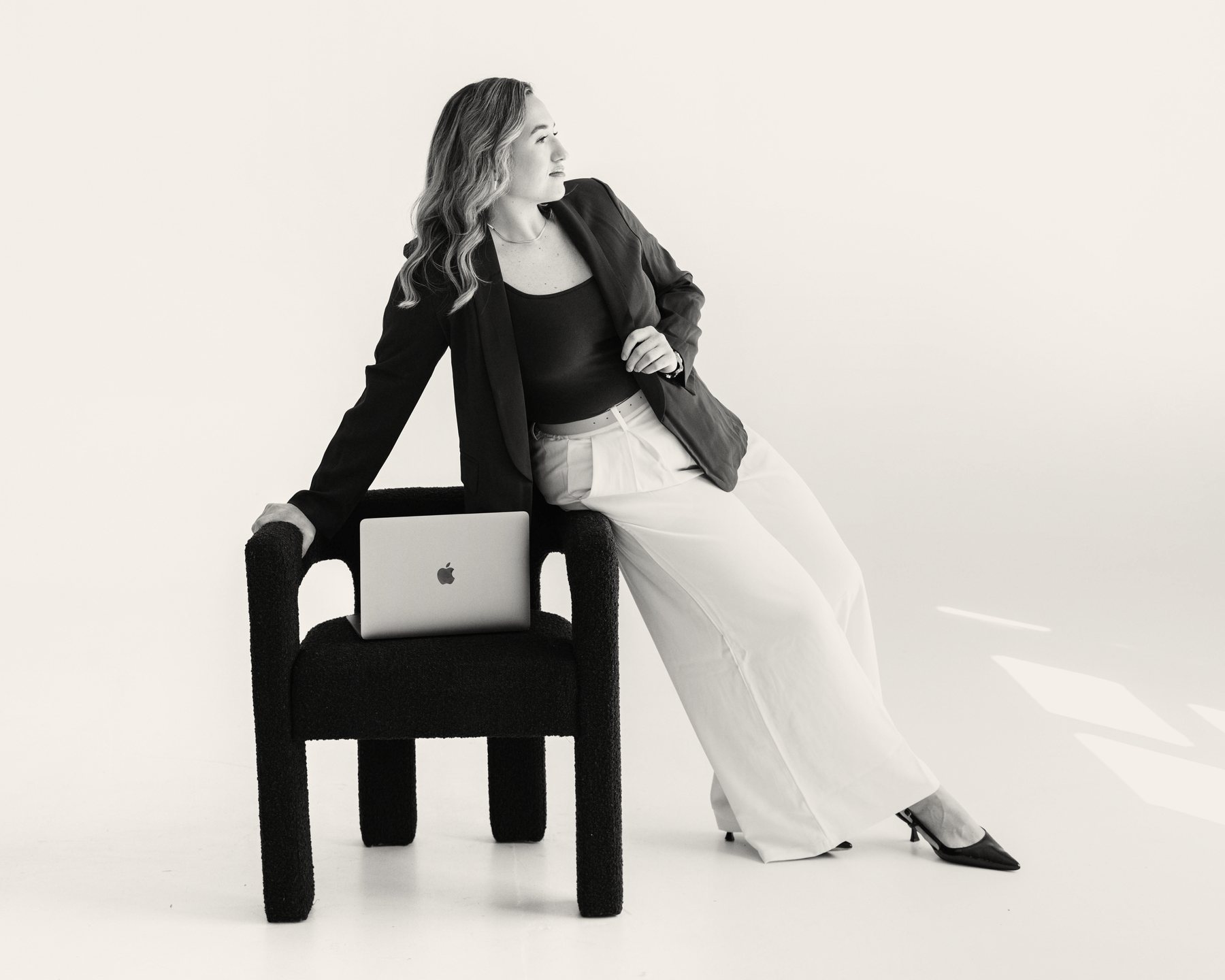 Kristin Pitts, founder of Aligned Consulting, seated on the arm of a chair in a white studio with a laptop beside her